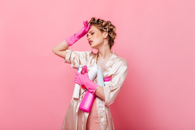 La mujer está cansada después de limpiar y posa con detergentes en la pared rosa