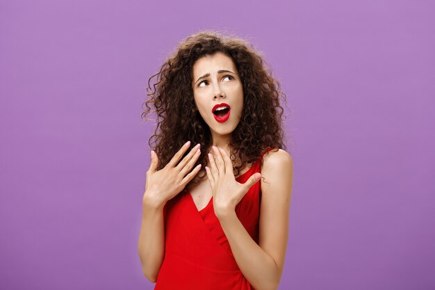 La mujer está complacida con su propia mirada enamorada de sí misma. Arrogante, sexy y elegante mujer de pelo rizado en elegante vestido de noche rojo saludando con las palmas como si se sintiera caliente mirando en la esquina superior derecha.