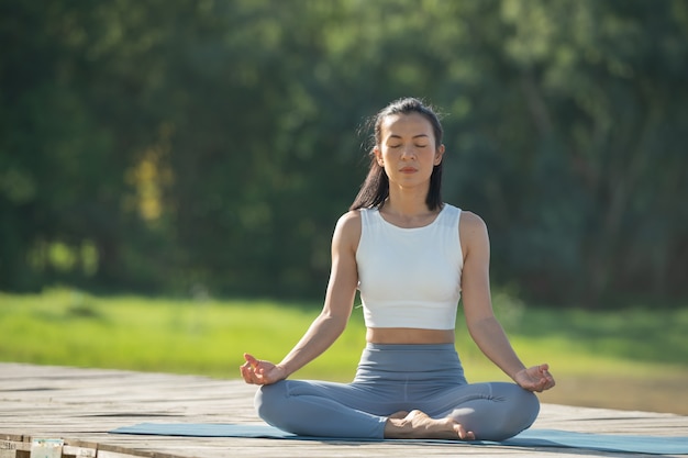Mujer en una estera de yoga para relajarse en el parque en el lago de la montaña. Mujer tranquila con los ojos cerrados practicando yoga, sentada en la pose de Padmasana en la estera, ejercicio de loto, atractiva chica deportiva en ropa deportiva.