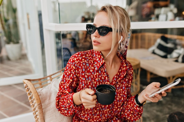 Mujer con estilo atractivo en gafas de sol negras tomando café y hablando por teléfono inteligente