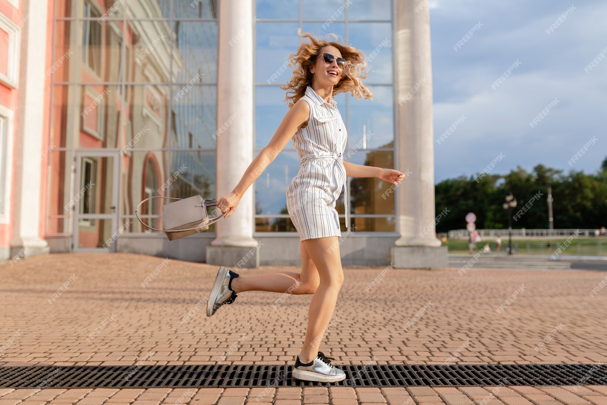 Mujer estilo atractivo joven corriendo saltando divertido en zapatillas de deporte en la calle de la ciudad en vestido de estilo de moda verano con gafas de sol y