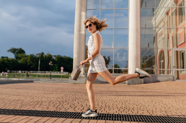 Mujer con estilo atractivo joven corriendo saltando divertido en zapatillas de deporte en la calle de la ciudad en vestido blanco de estilo de moda de verano con gafas de sol y bolso