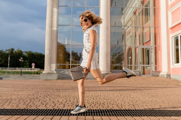 Mujer con estilo atractivo joven corriendo saltando divertido en zapatillas de deporte en la calle de la ciudad en vestido blanco de estilo de moda de verano con gafas de sol y bolso