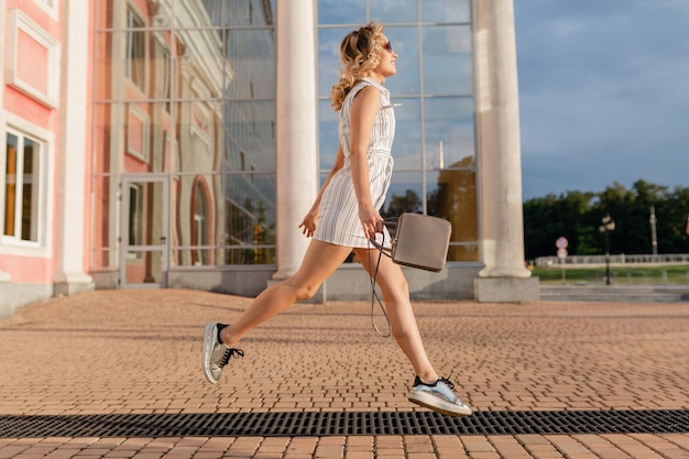 Mujer con estilo atractivo joven corriendo saltando divertido en zapatillas de deporte en la calle de la ciudad en vestido blanco de estilo de moda de verano con gafas de sol y bolso