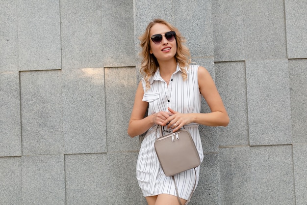 Mujer con estilo atractivo joven con peinado rizado rubio caminando en las calles de la ciudad en estilo de moda de verano vestido de rayas blancas con gafas de sol sosteniendo el bolso