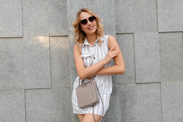 Mujer con estilo atractivo joven con peinado rizado rubio caminando en las calles de la ciudad en estilo de moda de verano vestido de rayas blancas con gafas de sol sosteniendo el bolso