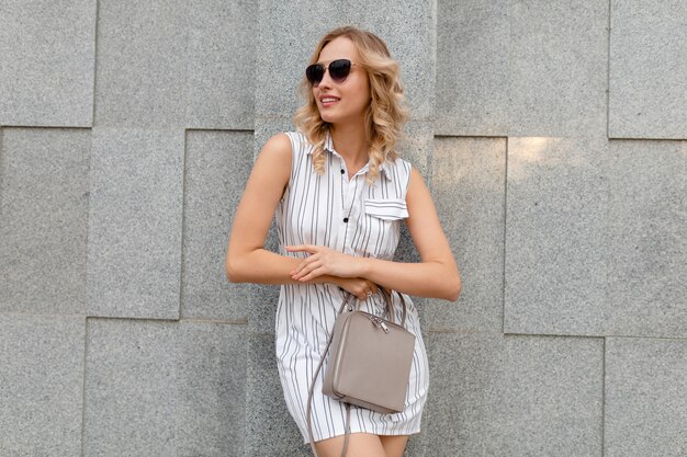 Mujer con estilo atractivo joven con peinado rizado rubio caminando en las calles de la ciudad en estilo de moda de verano vestido de rayas blancas con gafas de sol sosteniendo el bolso