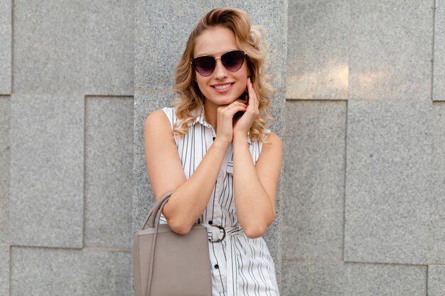 Mujer con estilo atractivo joven con peinado rizado rubio caminando en las calles de la ciudad en estilo de moda de verano vestido de rayas blancas con gafas de sol sosteniendo el bolso