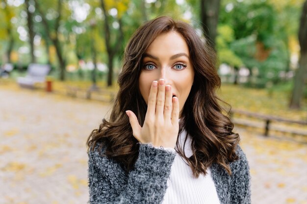 Mujer de estilo bonito está posando a la cámara en el parque con grandes emociones reales. Ella se ve sorprendida y cubre su rostro con su mano y muestra emociones reales.