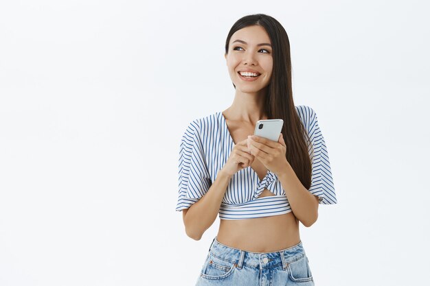 Mujer con estilo creativo en la parte superior recortada de moda mirando a la izquierda con una sonrisa encantadora sosteniendo el teléfono inteligente