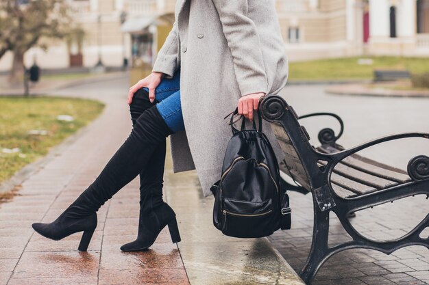 Mujer con estilo joven caminando en la ciudad de otoño, estación fría, con botas negras de tacón alto, mochila de cuero, accesorios, abrigo gris, sentado en el banco, tendencia de moda, detalles de primer plano de las piernas