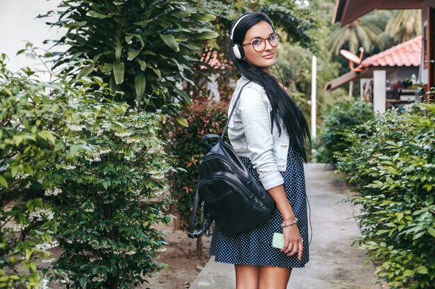 Mujer con estilo joven caminando, escuchando música en auriculares, sonriendo, feliz, sosteniendo la mochila, vacaciones de verano