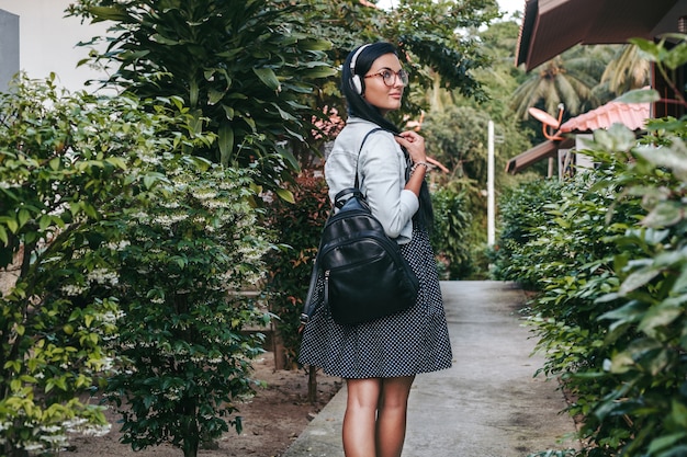 Mujer con estilo joven caminando, escuchando música en auriculares, sonriendo, feliz, sosteniendo la mochila, vacaciones de verano