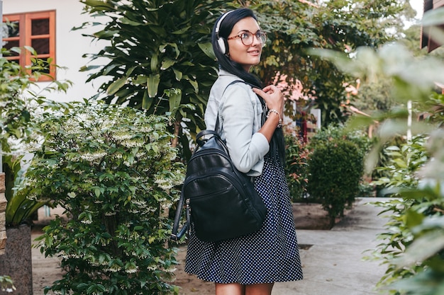 Mujer con estilo joven caminando, escuchando música en auriculares, sonriendo, feliz, sosteniendo la mochila, vacaciones de verano