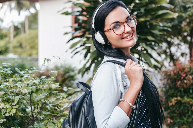 Mujer con estilo joven caminando, escuchando música en auriculares, sonriendo, feliz, sosteniendo la mochila, vacaciones de verano