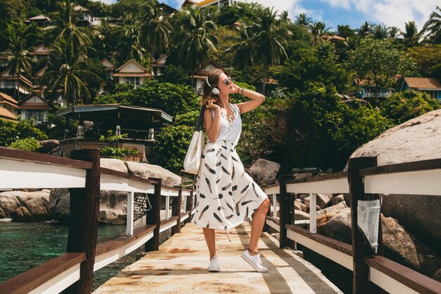 Mujer con estilo joven de pie en el muelle, caminar, escuchar música en auriculares, ropa de verano, falda blanca, bolso, agua azul, fondo de paisaje, laguna tropical, vacaciones, viajar en asia