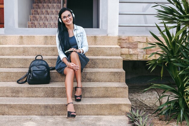 Mujer con estilo joven sentada en las escaleras con el teléfono inteligente, escuchando música con auriculares, mochila, sonriendo, estado de ánimo feliz y positivo, vacaciones de verano, estilo vintage denim