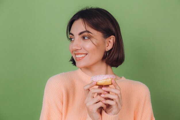 Mujer con estilo joven en suéter de melocotón casual aislado en la pared verde oliva con espacio de copia feliz rosquilla rosa