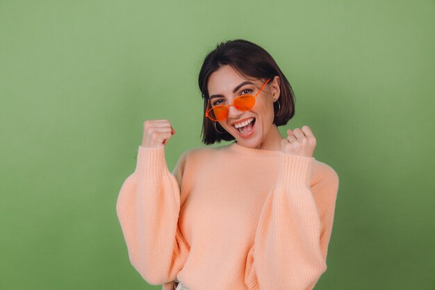 Mujer con estilo joven en suéter de melocotón casual y gafas naranjas aisladas en la pared de olivo verde positivo haciendo gesto ganador apretando espacio de copia de puño