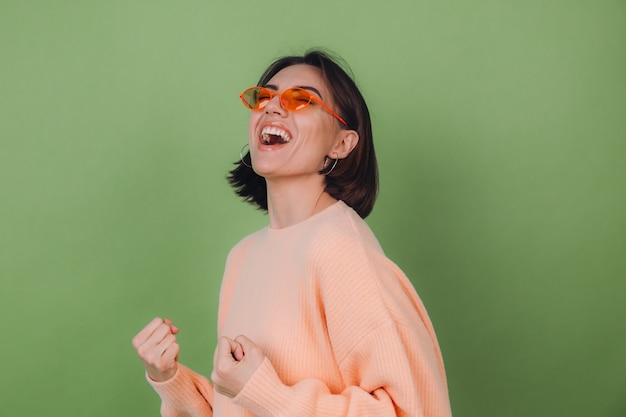 Mujer con estilo joven en suéter de melocotón casual y gafas naranjas aisladas en la pared de olivo verde positivo haciendo gesto ganador apretando espacio de copia de puño