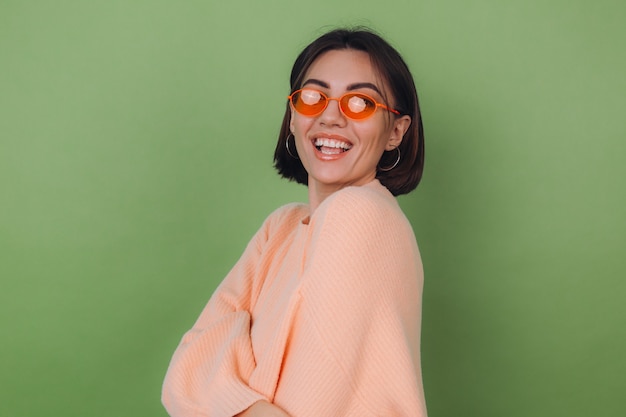 Mujer con estilo joven en suéter de melocotón casual y gafas naranjas aisladas en la pared de olivo verde risa sonriente positiva feliz alrededor de espacio de copia