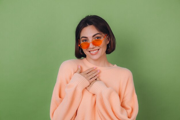 Mujer con estilo joven en suéter de melocotón casual y gafas naranjas aisladas en la pared verde oliva positiva tomados de la mano doblados en el pecho, espacio de copia de corazón