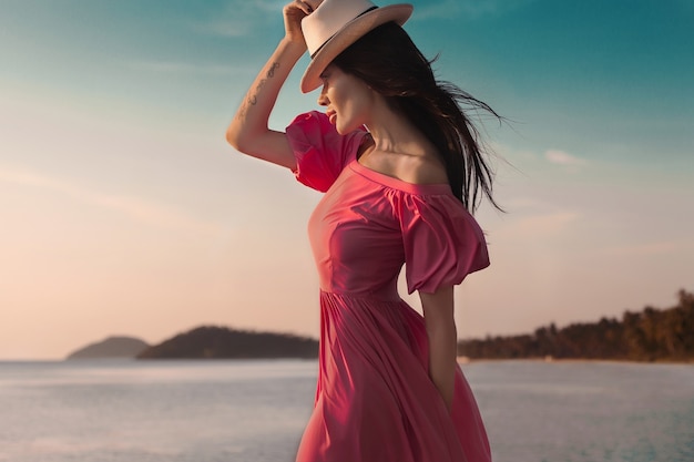 Mujer de estilo de verano al aire libre, retrato de moda soleado de mujer sensual con vestido rosa en la puesta de sol de la playa en la orilla del mar