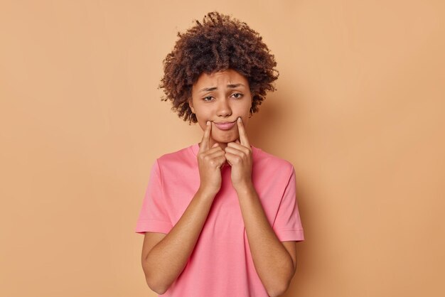 mujer estira la boca en una sonrisa forzada muestra su infelicidad viste una camiseta rosa casual aislada en la pared beige del estudio está triste sola. Concepto de emociones humanas negativas