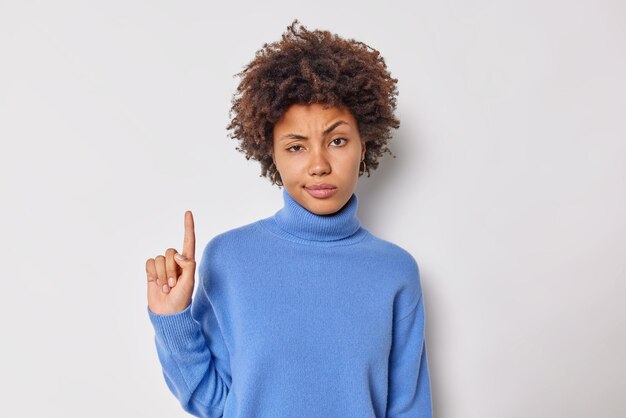 La mujer estricta, seria y atenta, señala con el dedo índice por encima de la cabeza, demuestra que algo en lo alto tiene una expresión de confianza en la cara y lleva un jersey azul casual aislado sobre fondo blanco. Solo mira esto