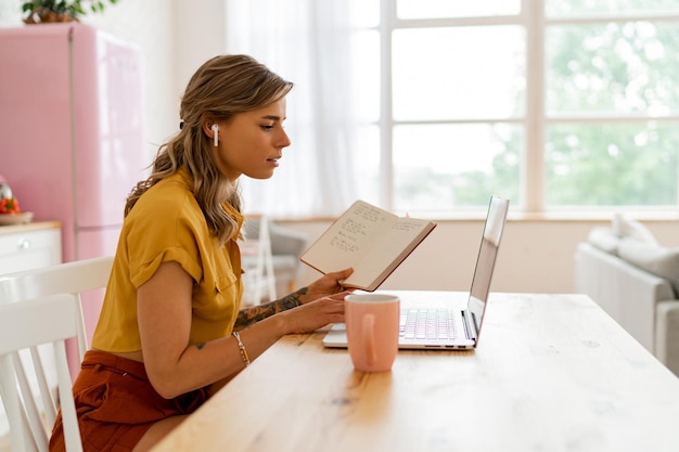 Mujer estudiante bonita que usa una computadora portátil y escribe notas en su cocina moderna y ligera