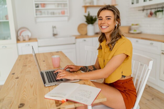 Mujer estudiante bonita que usa una computadora portátil y escribe notas en su cocina moderna y ligera