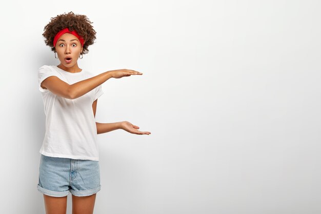 Mujer étnica fascinada explica el tamaño de algún objeto, habla de algo grande o grande, presenta un objeto enorme con sorpresa, mantiene la boca abierta, vestida con ropa informal, aislada en una pared blanca