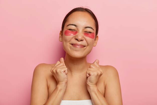 La mujer étnica llena de alegría cierra los puños con triunfo, disfruta de la rutina diaria de cuidados, cuida la piel, usa parches en los ojos, cierra los ojos por el placer recibido durante los tratamientos de belleza, posa en interiores