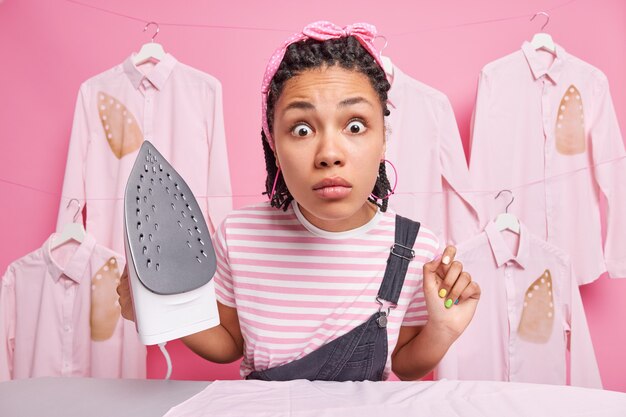 Mujer étnica muy sorprendida con trenzas mira fijamente sorprendida a la cámara ocupado planchando en casa sostiene plancha eléctrica vestida con ropa casual hace tareas domésticas diarias se opone a camisas planchadas