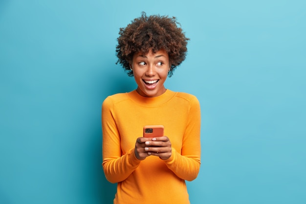 La mujer étnica de pelo rizado positivo usa el teléfono móvil, verifica los mensajes y lee las noticias. Tiene un teléfono celular moderno en las manos.