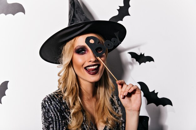 Mujer europea alegre posando juguetonamente en halloween. Adorable joven bruja con maquillaje negro que expresa felicidad.