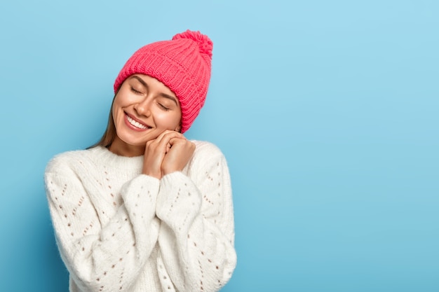 Una mujer europea alegre recuerda recuerdos agradables, inclina la cabeza, mantiene las manos cerca de la cara, sonríe alegremente, viste un gorro de punto rosado y un suéter blanco, posa sobre un fondo azul.