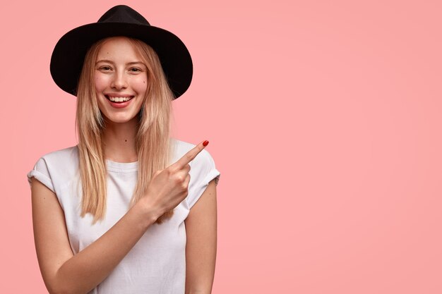 Mujer europea de aspecto agradable con apariencia atractiva, viste sombrero y camiseta informal, puntos a un lado