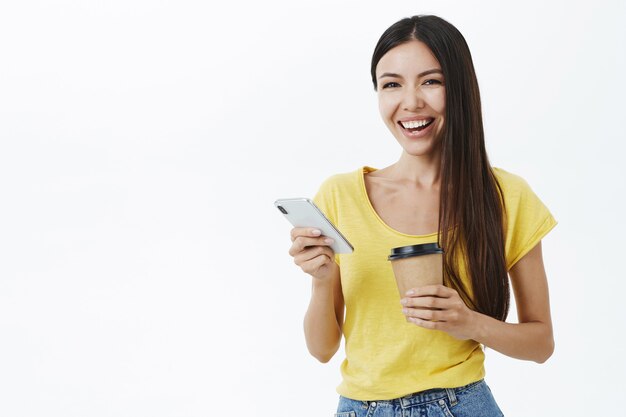 Mujer europea atractiva alegre y entretenida de aspecto amistoso con el pelo largo en la camiseta amarilla de moda que sostiene la taza de café de papel