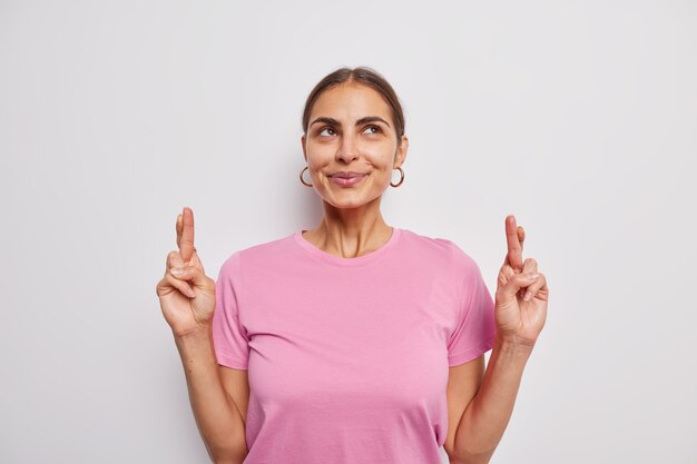 Mujer europea complacida mira hacia arriba rezando pide deseo con los dedos cruzados espera milagro espera resultados importantes viste camiseta rosa casual aislada sobre pared blanca