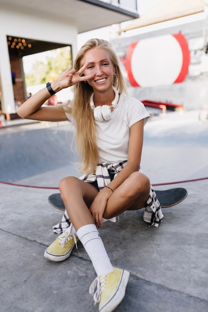 Mujer europea delgada posando con el signo de la paz después de patinar. Tiro al aire libre de magnífica dama bronceada viste camiseta blanca y auriculares grandes.