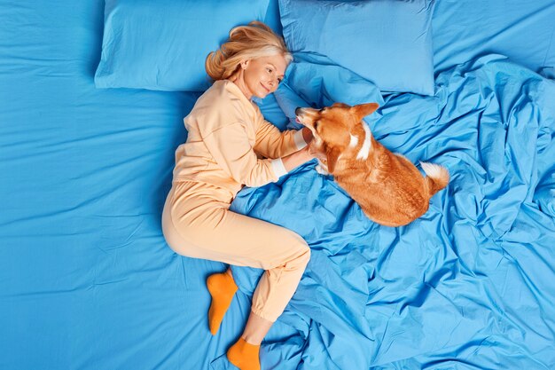 Mujer europea de edad complacida en ropa de dormir juega con su mascota favorita en casa en el dormitorio acostados juntos en la cama y disfrutar de un buen día. Mujer de mediana edad expresa amor y cuidado al perro como miembro de la familia