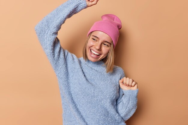 Una mujer europea feliz y optimista baila despreocupada, sacude los brazos, sonríe alegremente, mira hacia otro lado, tiene un estado de ánimo juguetón, usa un sombrero rosa y un saltador azul casual aislado sobre un fondo marrón, se mantiene siempre positivo