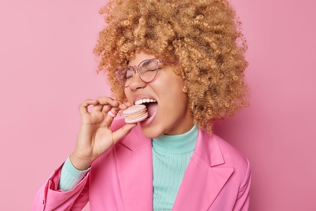 Mujer europea feliz con picaduras de pelo rizado apetitoso macarrón tiene adicción al azúcar vestida con ropa elegante usa gafas transparentes posa contra fundamento rosa Concepto de confitería