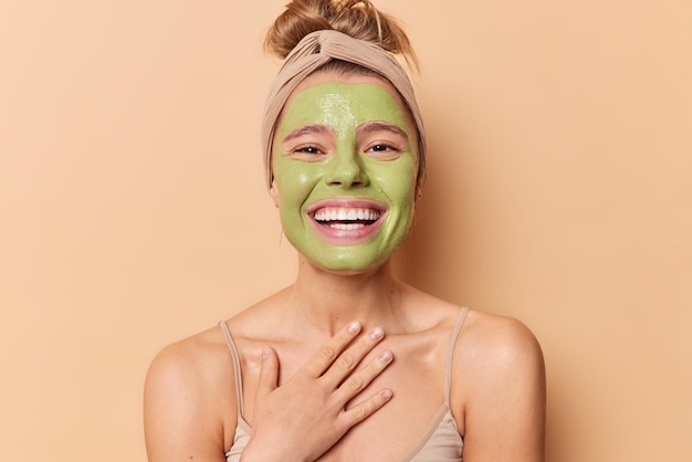 Foto gratuita la mujer europea feliz sonríe ampliamente, mantiene la mano en el pecho, aplica una máscara verde nutritiva, se somete a tratamientos de belleza, usa una diadema, tiene el pelo peinado aislado sobre un fondo marrón concepto de cuidado de la piel