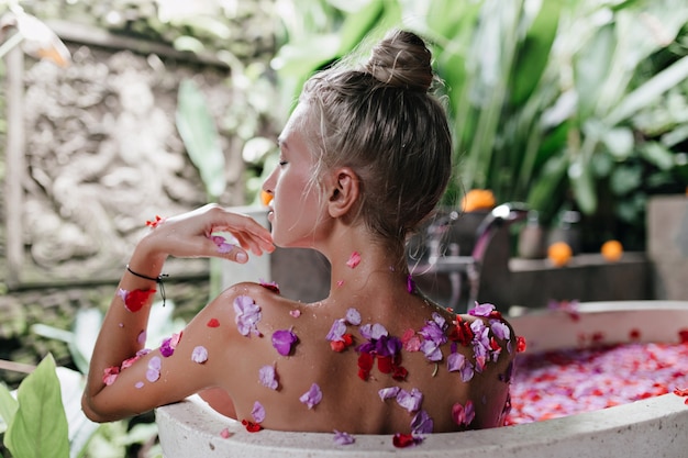 mujer europea haciendo spa con pétalos de flores en fin de semana. Retrato de interior de mujer alegre con piel bronceada relajante en el baño con expresión de cara feliz.