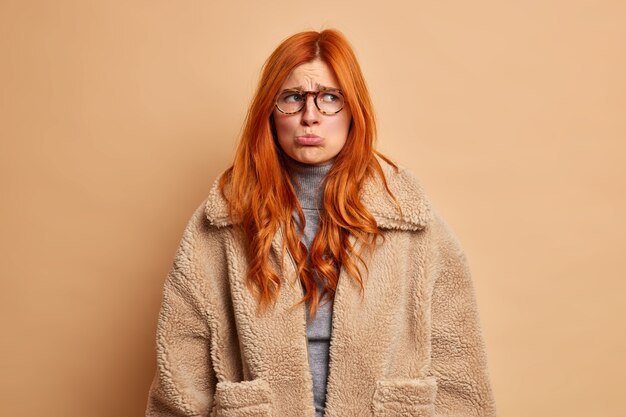 La mujer europea hosca ofendida con el pelo rojo frunce el labio inferior y mira a un lado con tristeza vestida con un abrigo de invierno marrón y quiere llorar de emociones desesperadas.