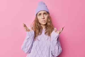 Foto gratuita una mujer europea indignada y desconcertada con el pelo ondulado se extiende las manos, muerde los labios, no sabe qué hacer, tiene una expresión despistada, lleva un suéter de punto y poses de sombrero sobre un fondo rosa. no me importa