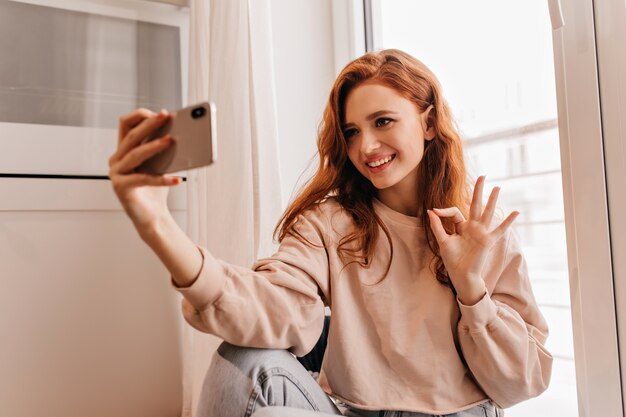 Mujer europea inspirada en ropa acogedora haciendo selfie. Retrato de interior de hermosa chica de jengibre.