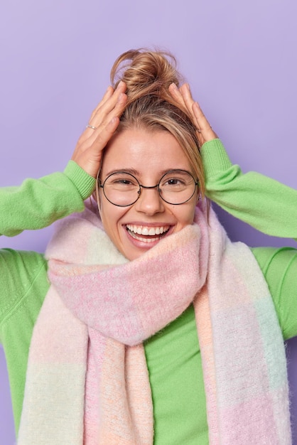 La mujer europea joven alegre agarra la cabeza siente felicidad sonríe ampliamente usa gafas redondas bufanda cálida alrededor del cuello se siente muy feliz mira directamente a la cámara aislada sobre fondo púrpura.